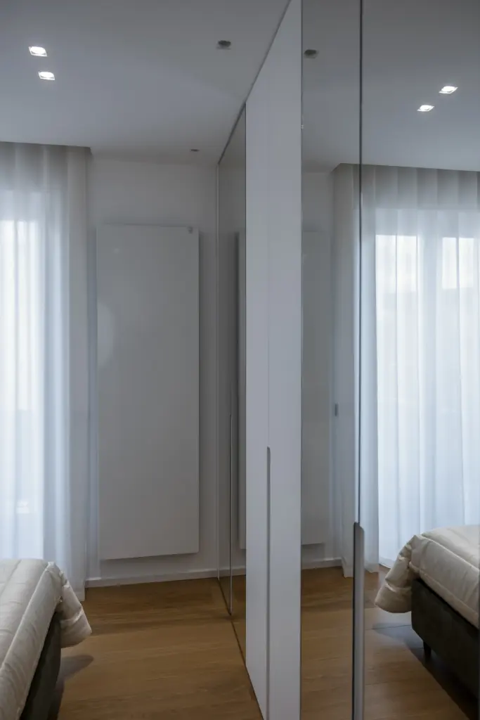 Interior minimalista con armadio bianco a tutta altezza con ante a specchio, tende bianche semitrasparenti e pavimento in parquet chiaro.