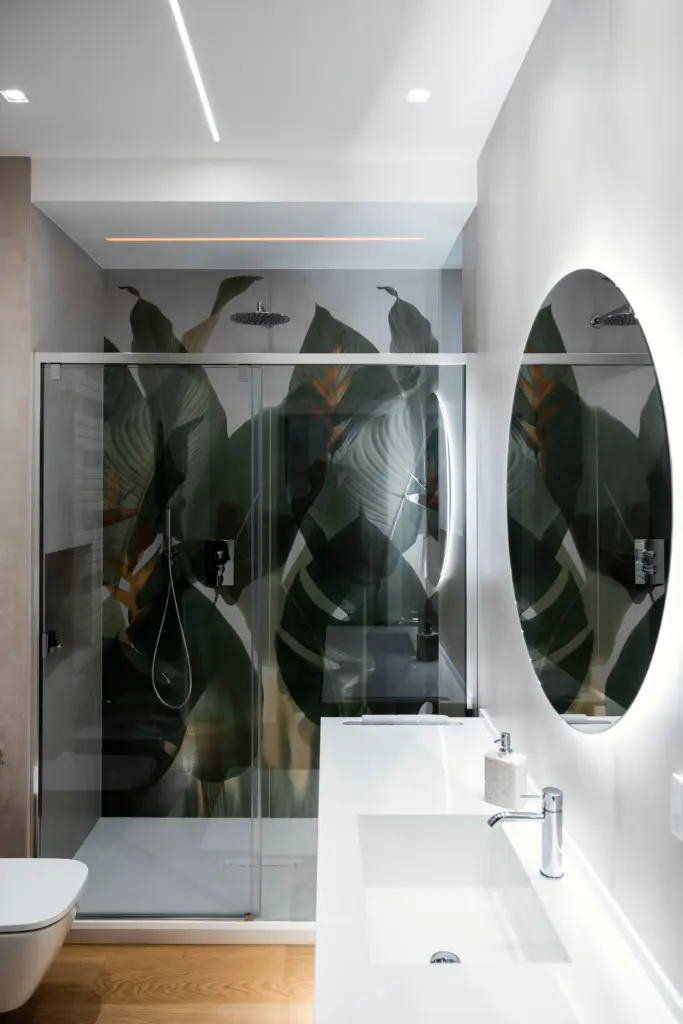 Bagno contemporaneo con grande specchio tondo, doccia in vetro e parete decorata con foglie tropicali