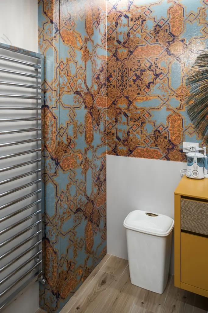 Dettaglio di un bagno moderno con rivestimento a mosaico geometrico in tonalità arancioni e azzurre, pavimento in legno e arredi giallo ocra.