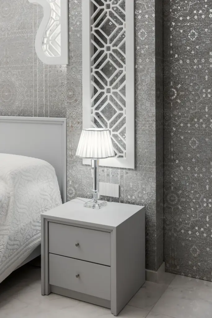 Camera da letto moderna con comodino grigio chiaro, lampada in cristallo con paralume plissettato bianco, specchio decorativo geometrico e carta da parati con motivi orientali.