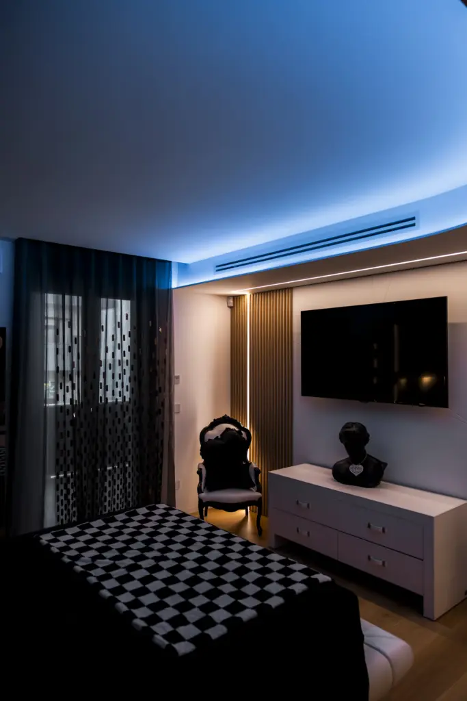 Camera da letto moderna con luci LED blu a soffitto, TV a parete e arredamento elegante.