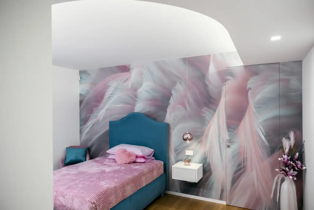 Camera da letto moderna per ragazza, con parete decorata a tema piume, letto imbottito azzurro e dettagli rosa.