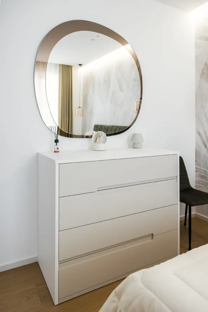 Cassettiera moderna bianca con linee minimaliste, accompagnata da uno specchio ovale di design e decorazioni delicate.