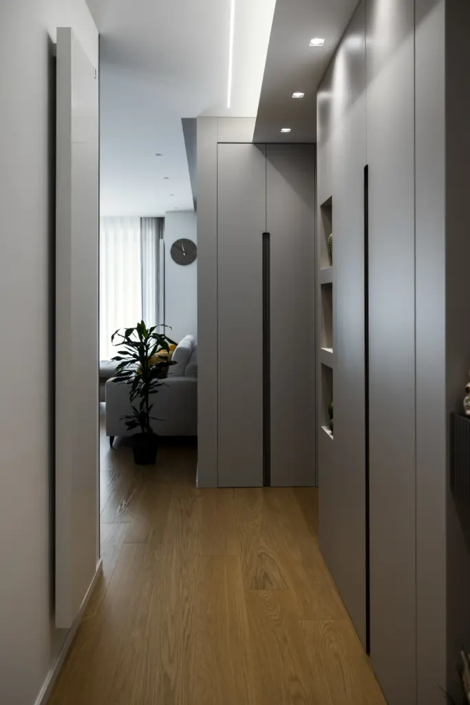 Corridoio elegante con pareti bianche, armadio integrato con maniglie verticali, pavimento in parquet e vista sul soggiorno con divano chiaro.