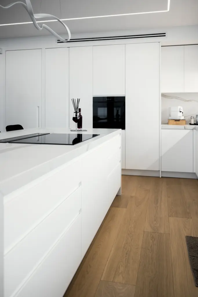 Cucina moderna bianca con isola centrale, piano a induzione e pavimento in legno naturale.