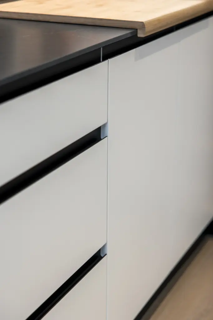 Dettaglio di cassetti di cucina moderna bianchi opachi con piano in marmo nero e tagliere in legno naturale.