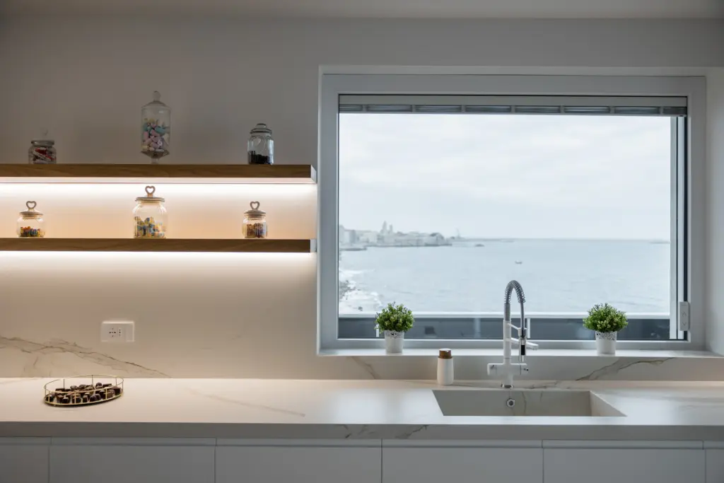 Cucina bianca moderna con piano effetto marmo, mensole retroilluminate e grande finestra con vista sul mare.