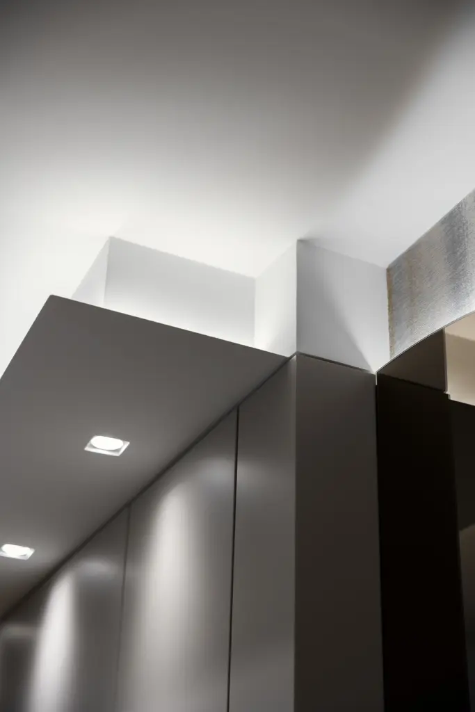 Dettaglio minimalista dell'angolo tra armadio e controsoffitto con faretti LED incassati e giochi di luce su superfici bianche e grigie.