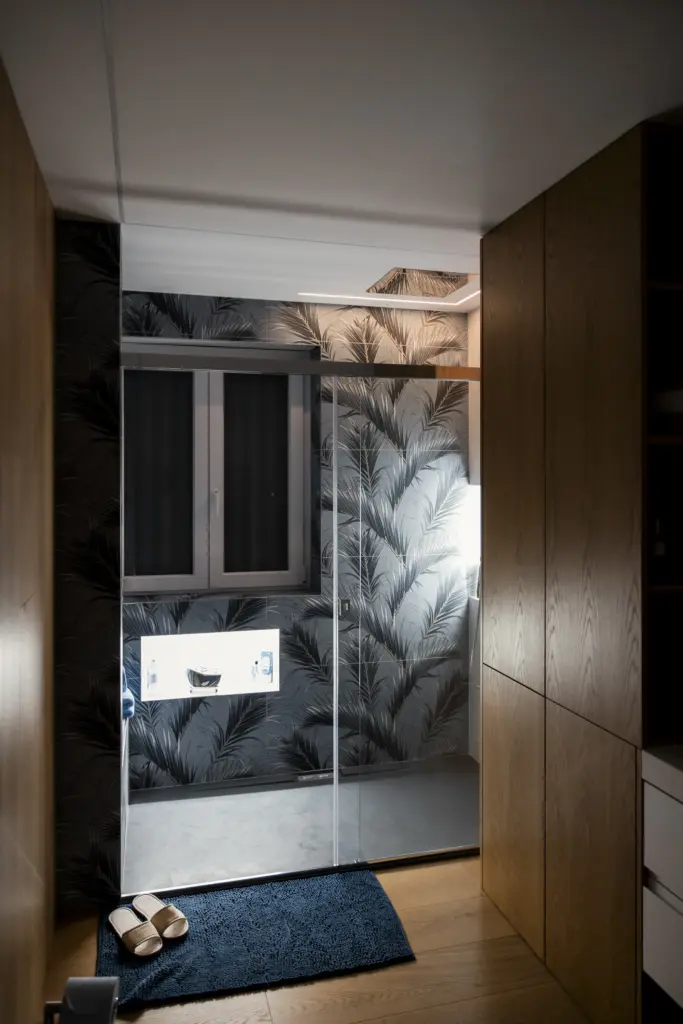 Bagno moderno con parete decorata con motivo foglie nere, cabina doccia in vetro e arredo in legno.
