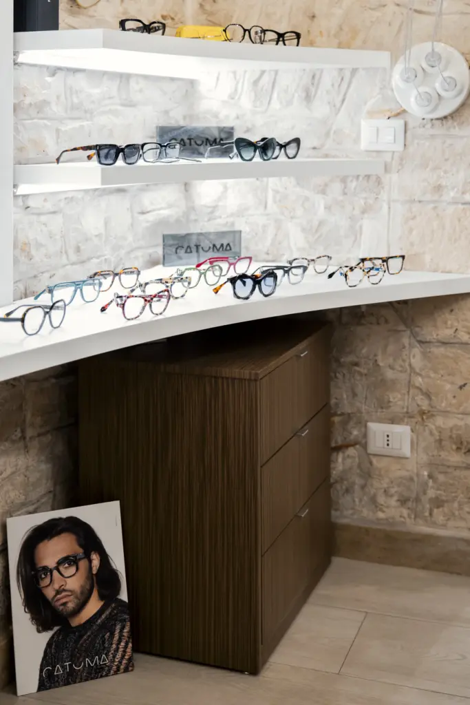 Ripiani angolari bianchi per esposizione di occhiali da vista con cassettiera in legno zebrato e sfondo in pietra.