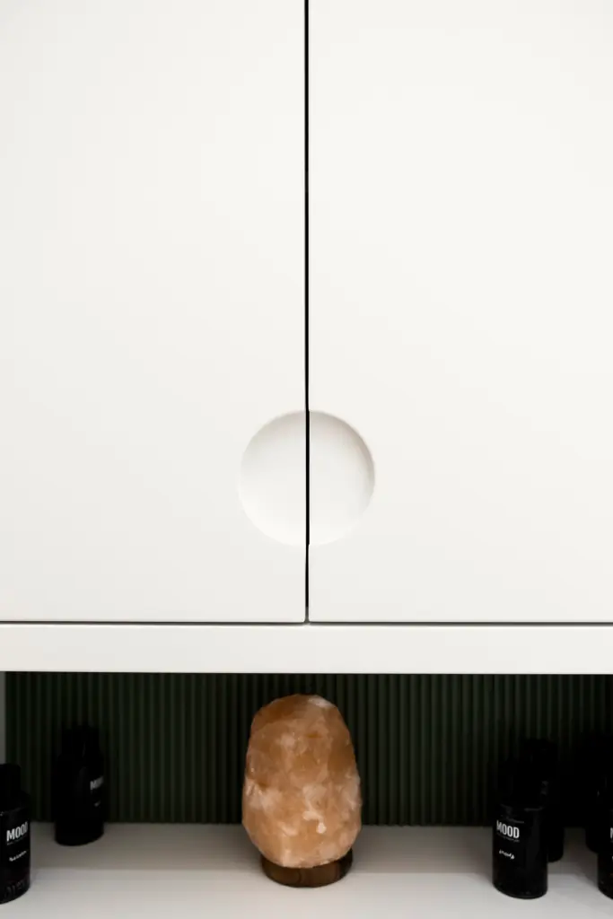 Mobile contenitore bianco con maniglie scavate a mezzaluna, retro con pannello verde cannettato e lampada di sale decorativa