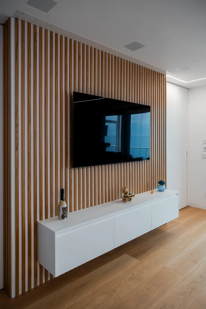 Parete attrezzata con pannelli verticali in legno chiaro, mobile bianco sospeso e TV a schermo piatto installata centralmente