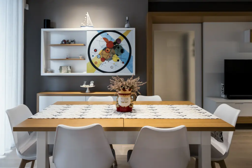 Sala da pranzo moderna con tavolo in legno chiaro, sedie bianche e arredo minimalista su sfondo grigio.