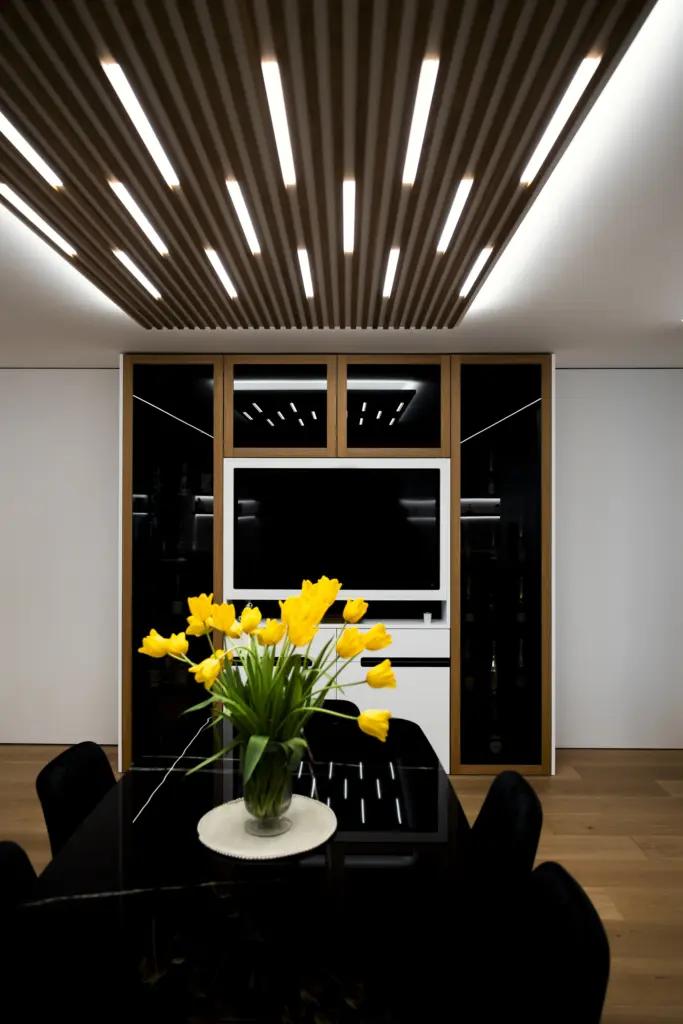 Zona pranzo con tavolo nero lucido, tulipani gialli, soffitto in doghe di legno e illuminazione LED integrata