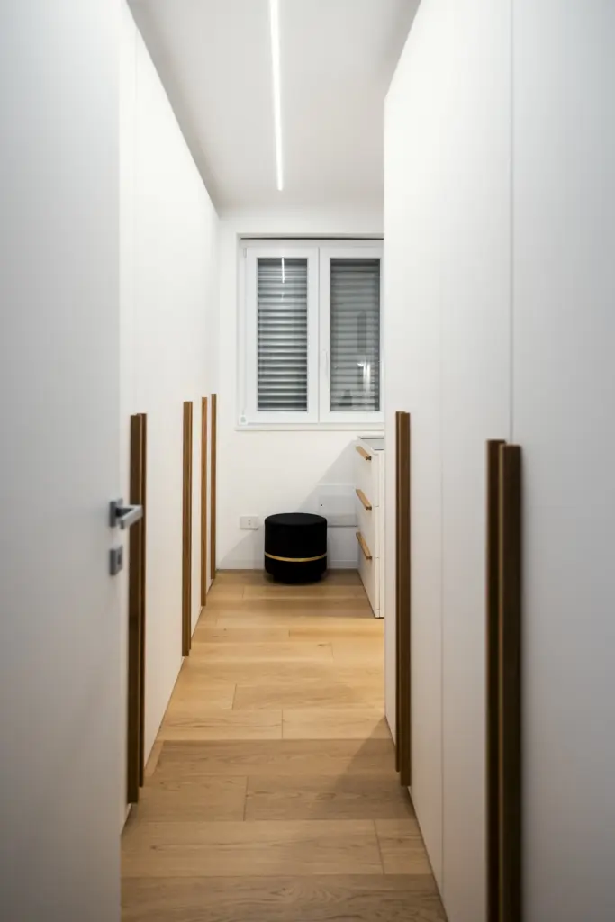Vista interna di una cabina armadio moderna con armadi bianchi e maniglie verticali in legno naturale.